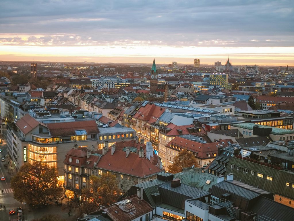 Ein Blick auf die Häuser von München von oben.
Im Hintergrund ein bunter Abendhimmel mit Wolkenschicht.