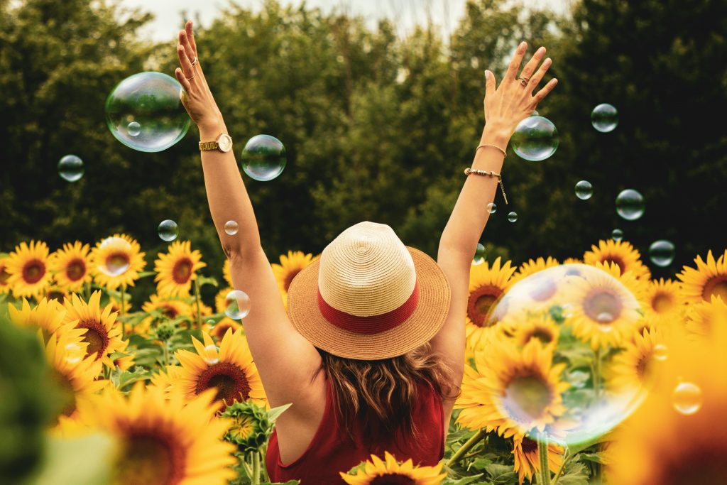 eine Frau mit Hut steht in einer Wiese mit Sonnenblumen und streckt vor Freude die Arme in die Höhe; um sie herum fliegen Seifenblasen