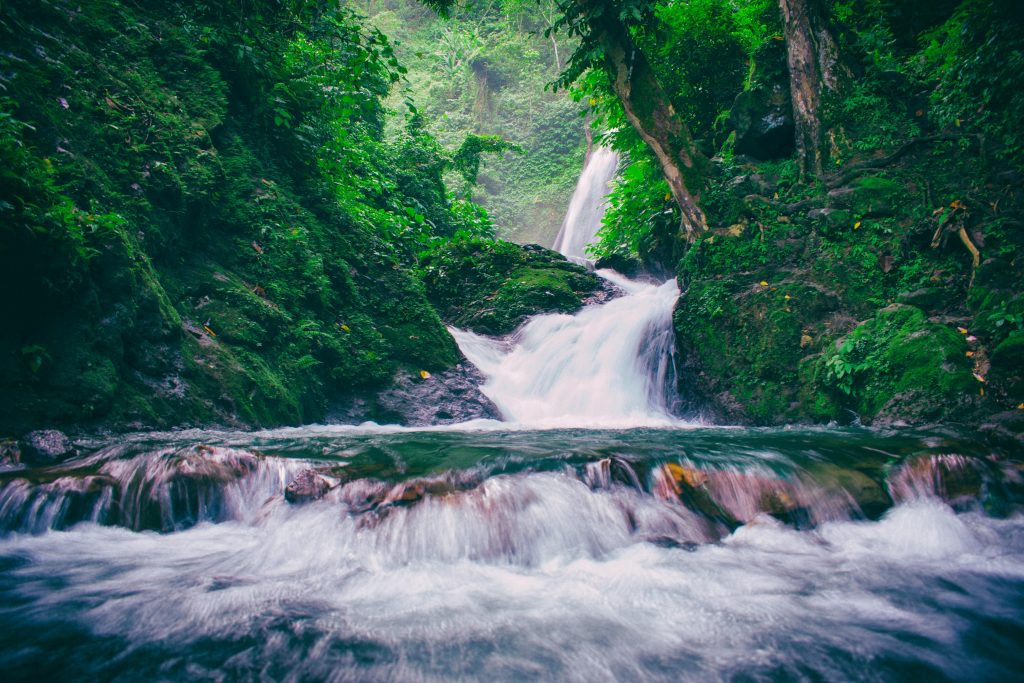 Durch moosbewachsene Felsen fließt ein Fluss.
An manchen Stellen ist er ein kleiner Wasserfall.