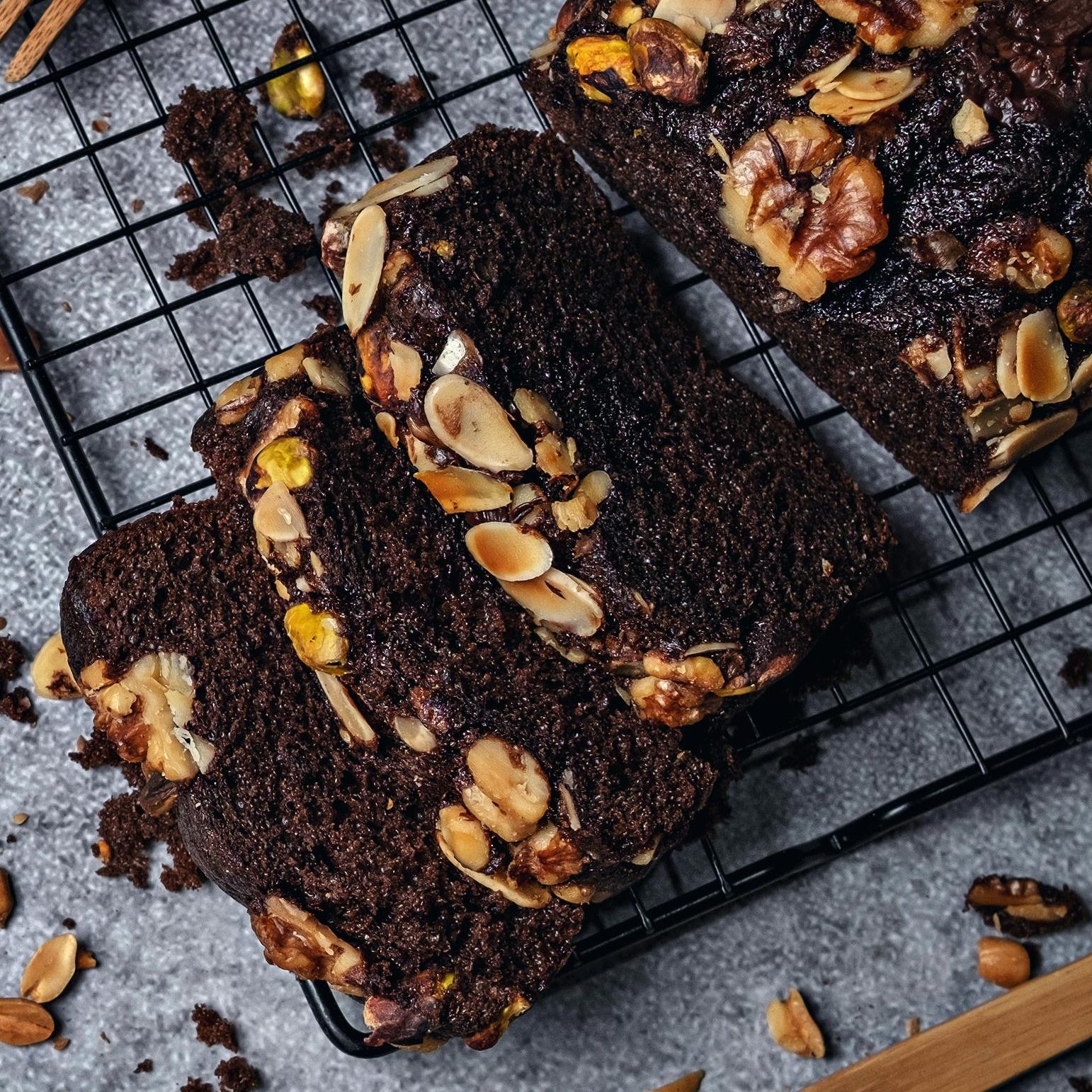 Auf einem Ofengitter steht ein dunkelbrauner Schokoladenkuchen mit Walnüssen oben drauf.
Drei Stücke sind abgeschnitten und liegen davor.