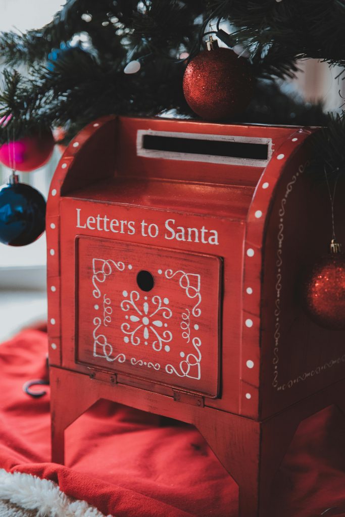Auf einem roten Teppich steht ein roter Behälter mit der Aufschrift "Briefe an den Weihnachtsmann".
Darüber sieht man Teile eines Christbaums mit roten, rosanen und blauen Christbaumkugeln.