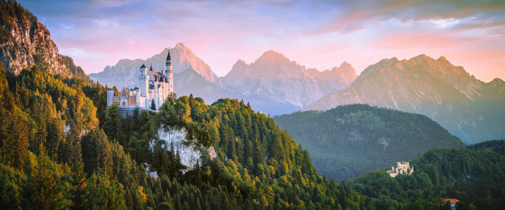 Von der Ferne sieht man Schloss Neuschwansten, wie es auf den Bergen tront.
Dahinter sieht man weitere majestätische Berge und darüber einen blau-grau-rosa gefärbten Himmel mit Wolken.