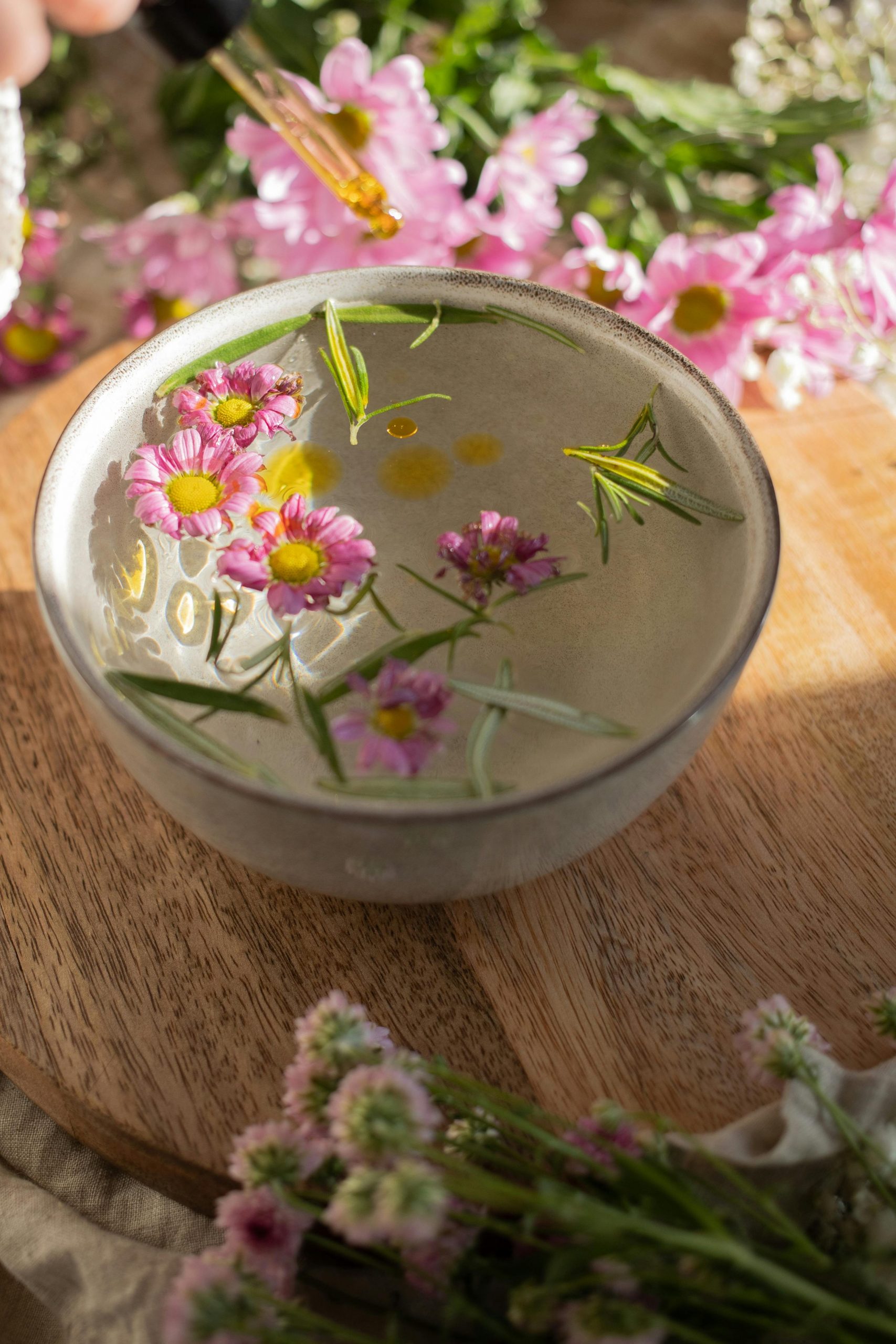 Auf einem Hoilztisch steht ein Schälchen mit Wasser.
Darinnen schwimmen rosarote Blüten und grüne Rosmarinnadeln.
Um das Schälchen herum befinden sich verschiedene Blumen.