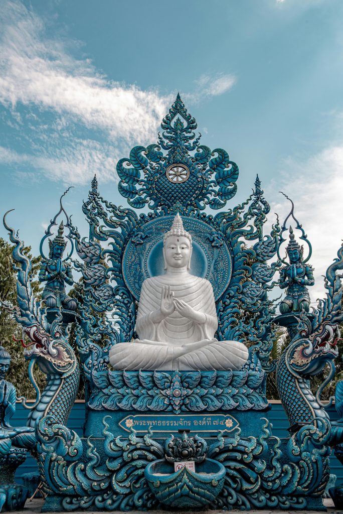 Gegen einen blauen Himmel ragt eine weiße Buddha-Statue im Lotussitz empor. Sie befindet sich auf einem prachtvollen, steinernen, reich verzierten, hellblauen Tron.