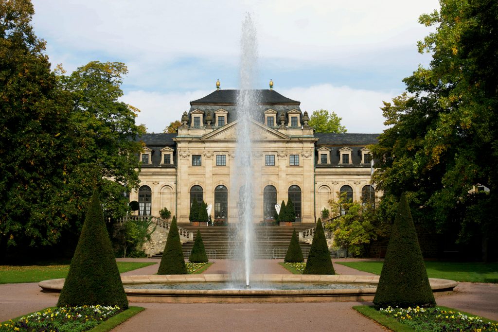 Ein Schloss. Daneben Bäume.
Davor eine Fontäne und kleine Bäumchen, die zugeschnitten sind, ie spitze Hütchen.
Alles ist symmetrisch angeordnet.