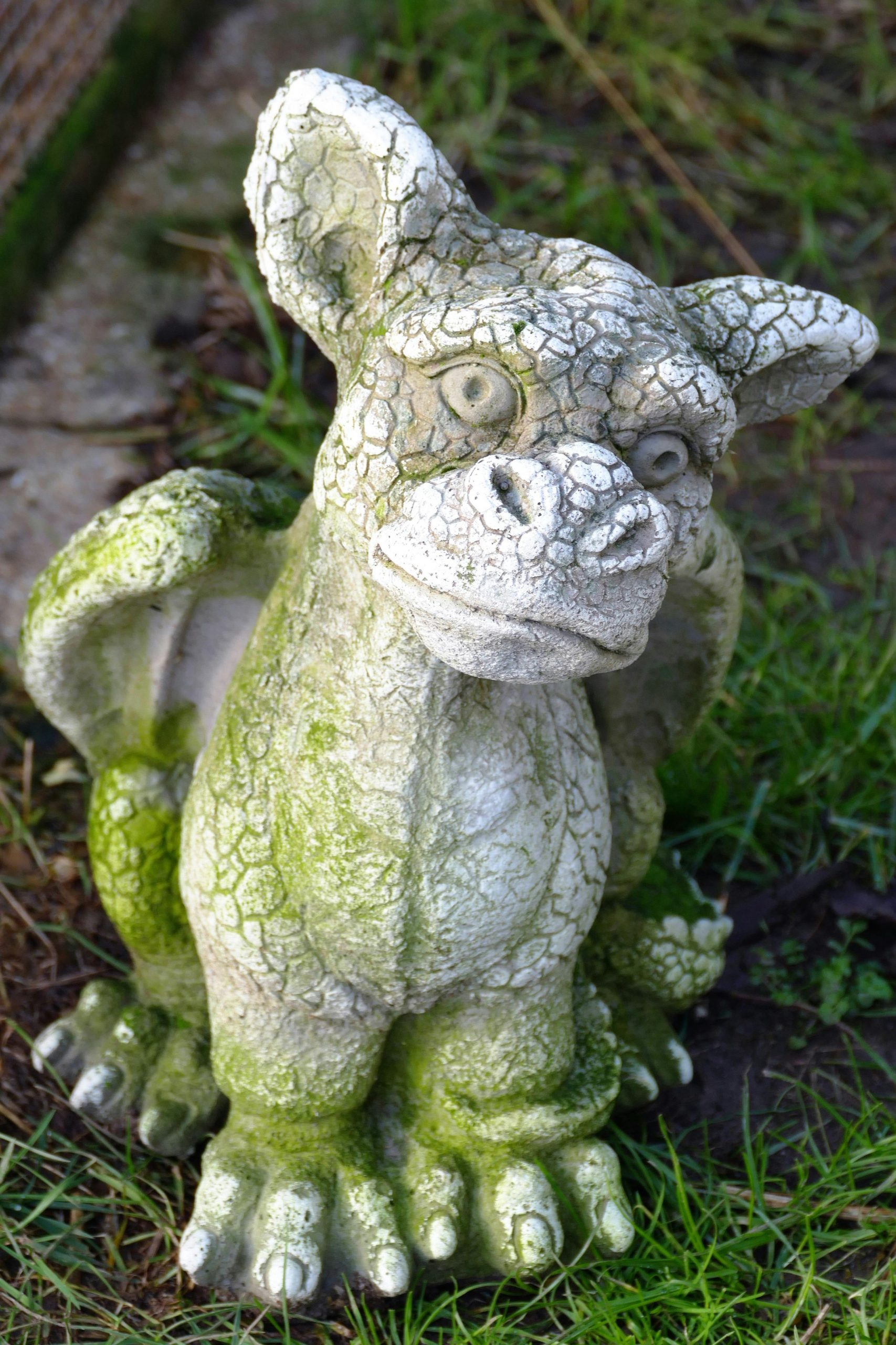 Auf Gras steht die große Figur eines kleinen, marmorfarbenen Drachen mit einem Hundeblick.
Auf ihm befindet sich an manchen Stellen Moos.