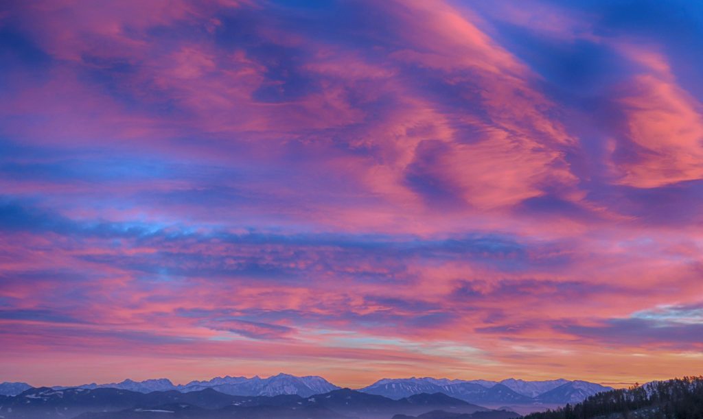 Ein bunter Himmel über schneebedeckten Bergen.
Die Farben sind: dunkelblau, hellblau, rosa, pimk, apricot.