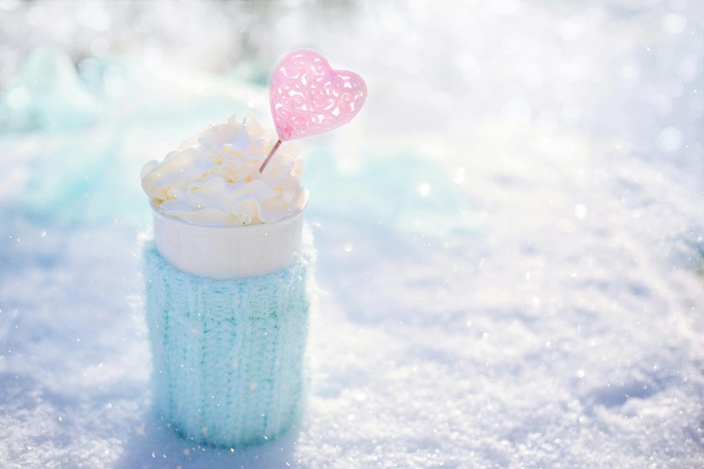 Auf in der Sonne glitzerndem Schnee steht ein Becher. Um ihn gewickelt ist ein hellblaues Strickmäntelchen.
Er hat eine große Haube aus Sahne.
Darin steckt ein Stöckchen mit einem rosanen Herz oben drauf.