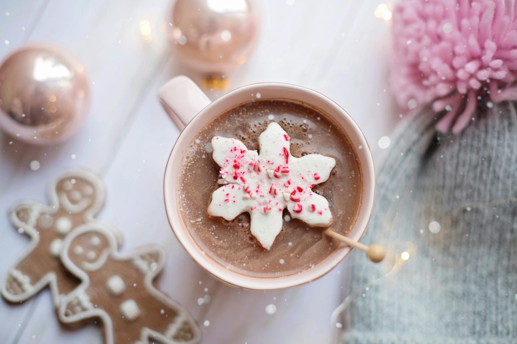 Auf einem weißen Holztisch steht eine Tasse. Darin befindet sich heiße Schokolade. Auf der Flüssigkeit liegt eine Süßigkeit in Form einer weißen Blume.
Um die Tasse herum stehen goldene Christbaumkugeln, eine graue Mütze mit rosanem Bommel und zwei Lebkuchenmännchen.