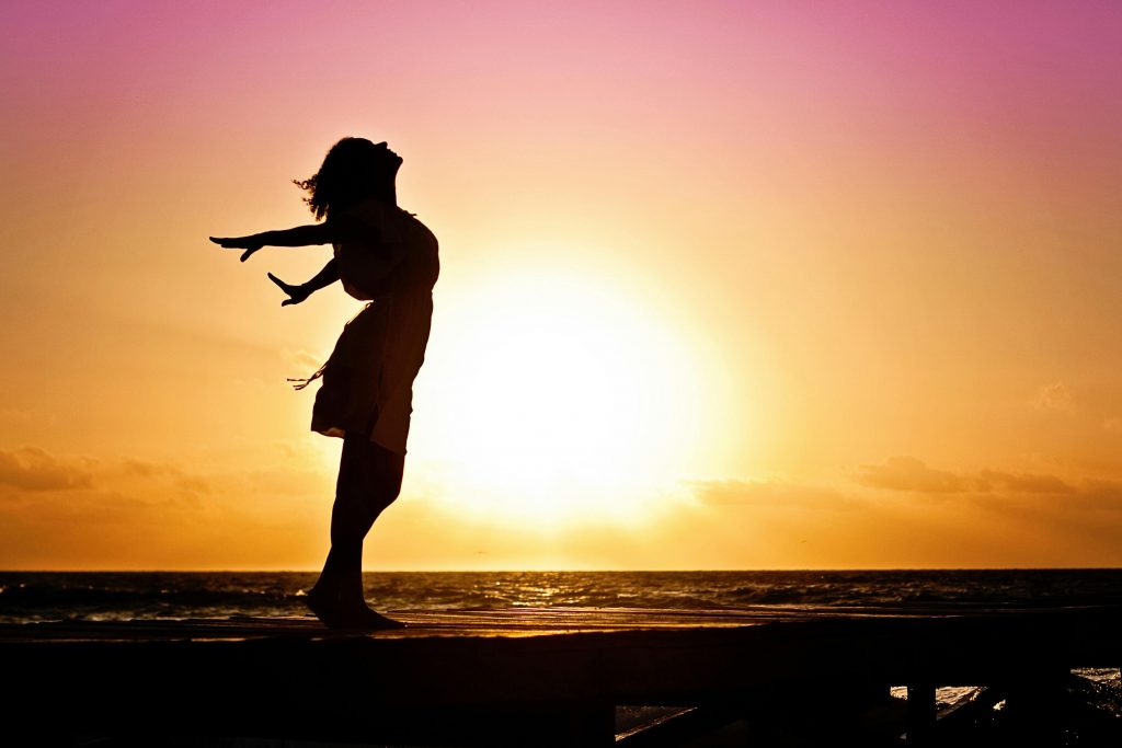 Eine Frau steht am Meer und streckt die Arme nach hinten. Hinter ihr ist die Sonne und ein farbenfroher Himmel.
grenzenlose Freiheit