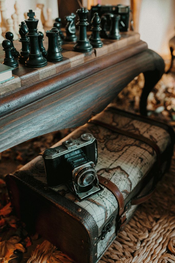 Auf einer alten Reisekiste steht eine alte Fotokamera.
Dahinter steht ein altes niedriges Tischchen aus dunkelbraunem Holz mit Schachfiguren drauf.