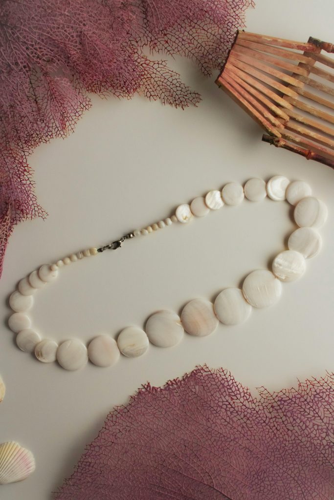 Auf einem weißen Untergrund liegt eine Kette aus runden, weißen Muschelscheibchen.
Als Deko wurden drum herum rosafarbene Korallenskelette drappiert.