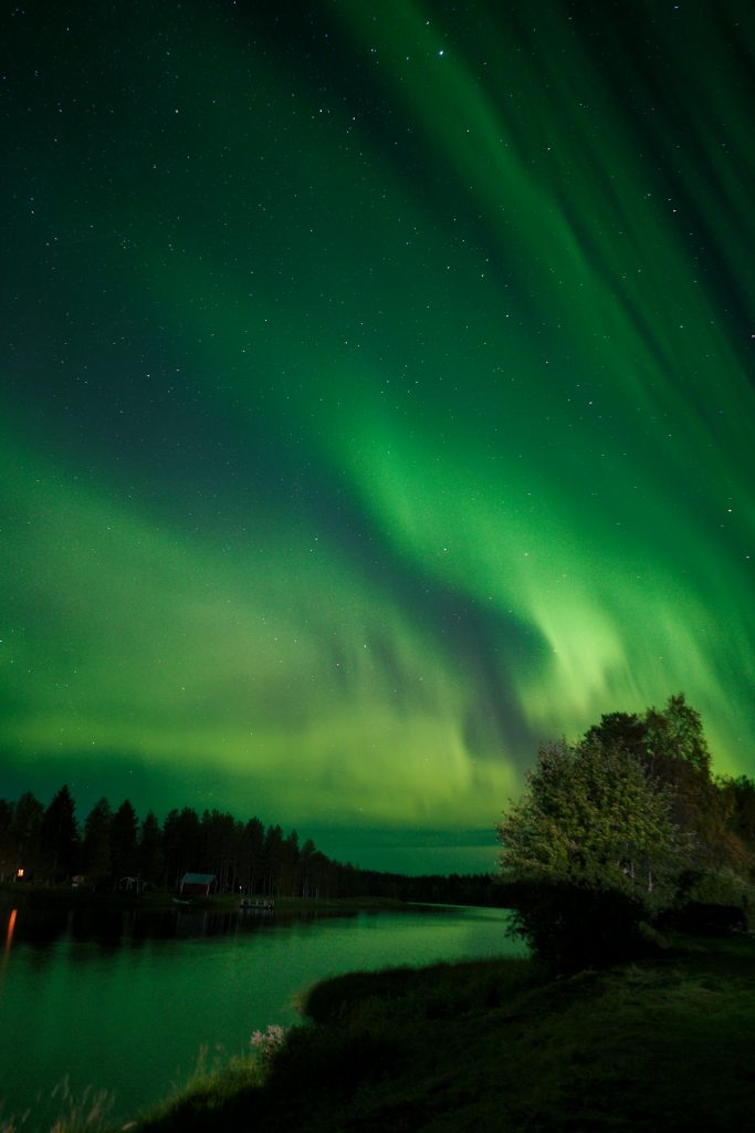 ein Fluss mit Bäumen
Darüber leuchtet am dunklen Himmel grünlich die Aurora Borealis.
