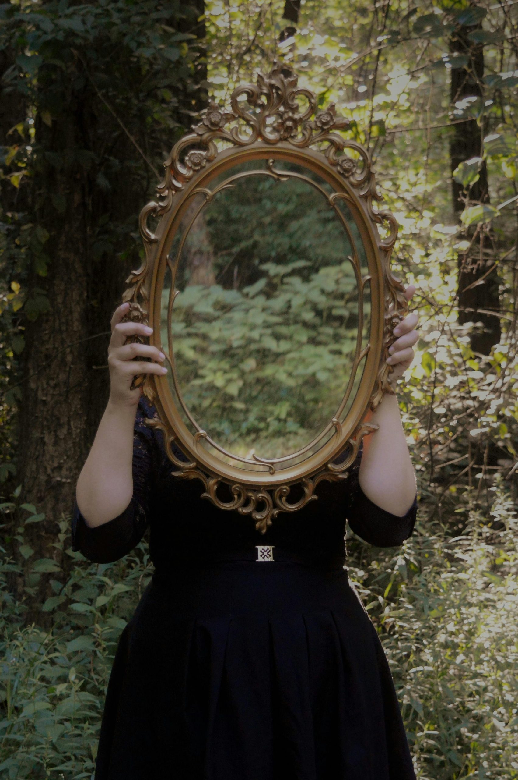 Eine Frau im schwarzen Kleid steht im Wald und hält einen ovalen, gold eingerahmten Spiegel vor Kopf und Oberkörper.
Im Spiegel spiegelt sich der Wald.