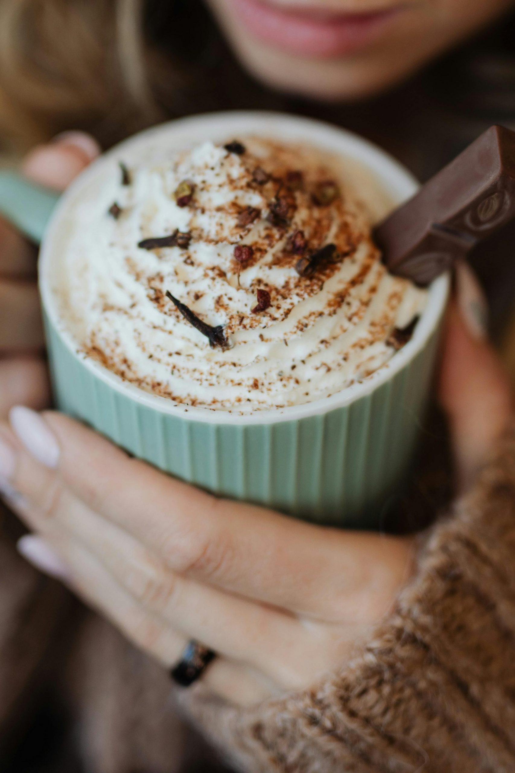 Eine Frauenhand mit braunem Pullover und rosanen Fingernägeln hält eine türkisfarbene Tasse in der Hand.
Darin befindet sich ein Häubchen Sahne mit Schokoflocken und Schokopulver.
In der Tasse steckt ein Schokoriegel.
Man sieht die leicht geöffneten Lippen der Frau.