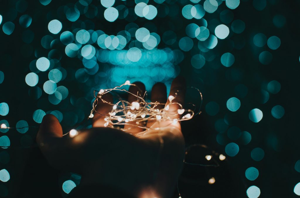 Es ist dunkel.
Auf einer Hand liegt eine Lichterkette, deren Lichter aus hellen Punkten bestehen.
Im Hintergrund sind verschwommene, türkise Kreise zu sehen.