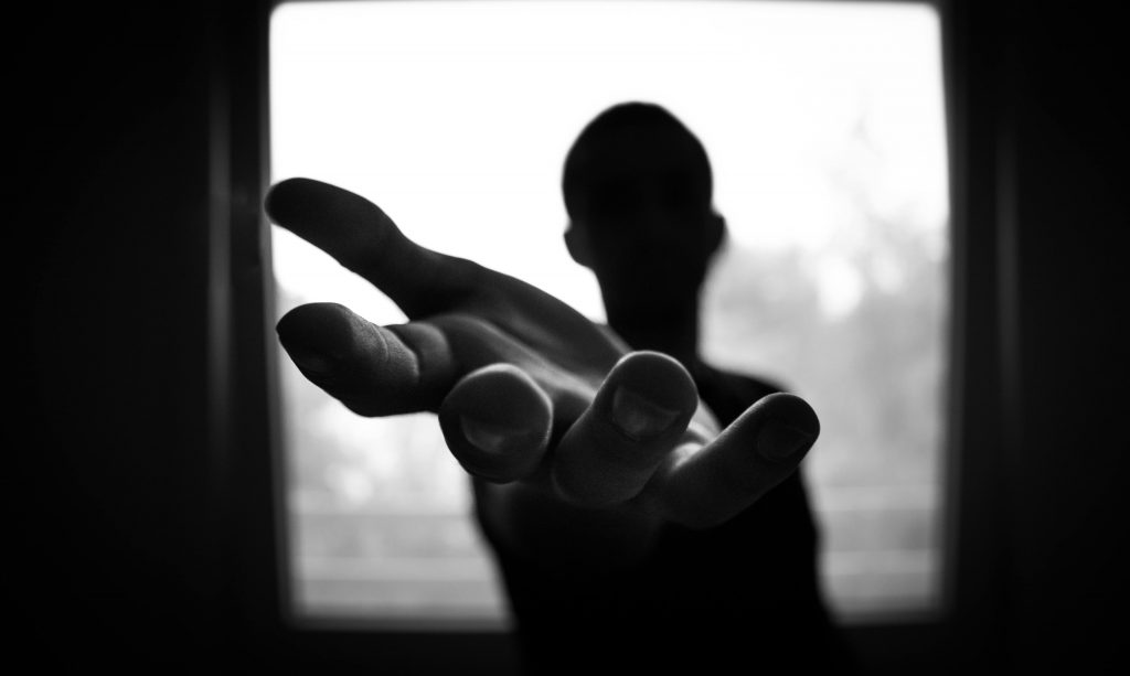 Die schwarze Silhouette eines Mannes steht vor einem Fenster.
Er streckt die Hand Richtung Kamera.