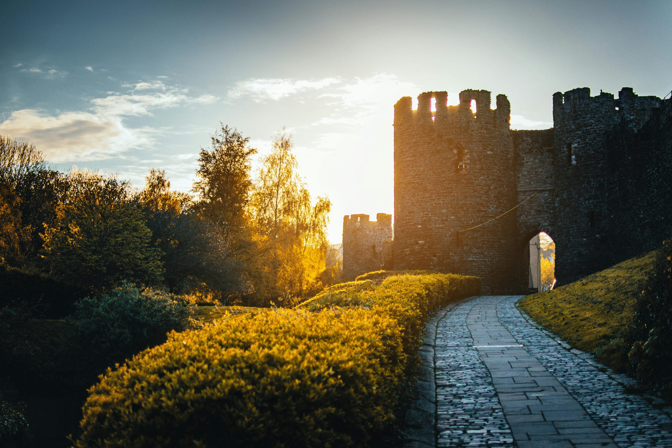 ein Weg führt in eine Burg, die Abendsonne blinzelt dahinter hervor