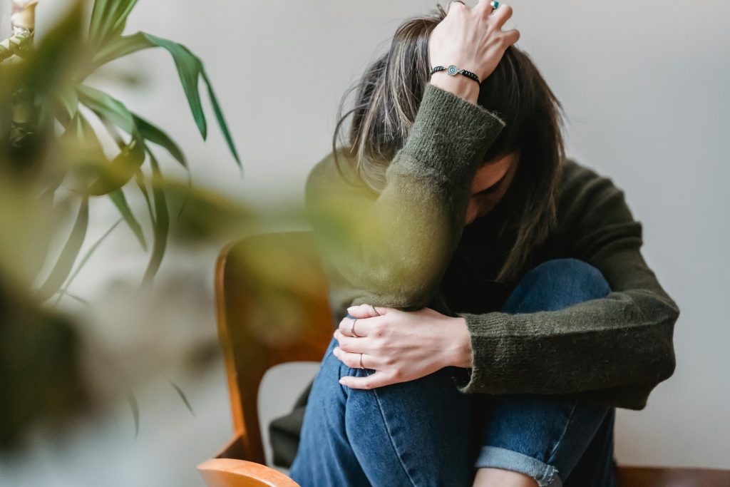 Eine Frau mit schwarzen Haaren sitzt auf einem Stuhl.
Ihre Beine sind angezogen.
Ihr Kopf ist darin vergraben.
Ein Arm hält ihre Beine, der andere den Kopf.