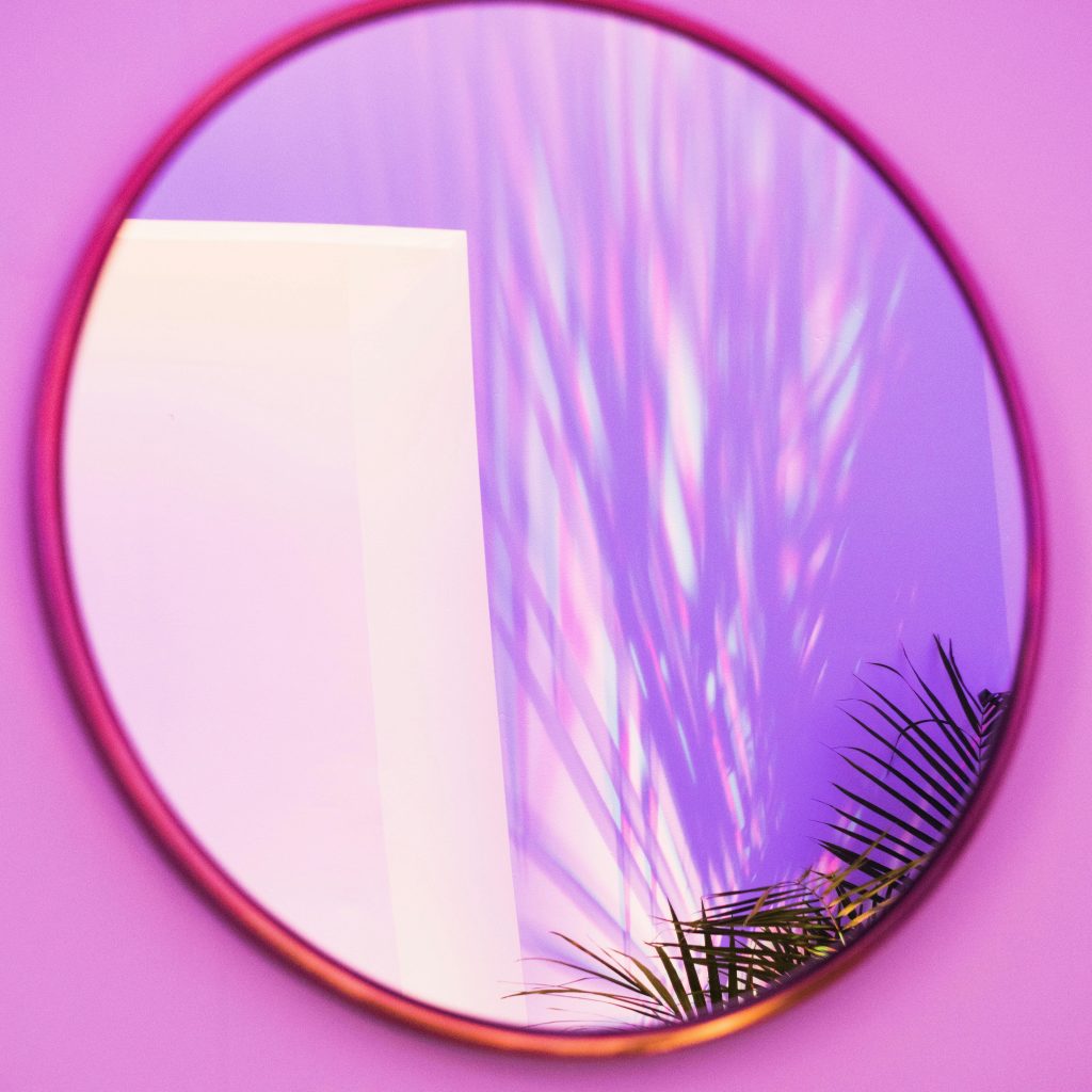Ein runder Spieghel mit goldenem Rahmen.
Die Wand im ihn ist pink-rosa.
In ihm sieht man die Spitzen einer Zimmerpalme und deren Schatten an der Wand.
