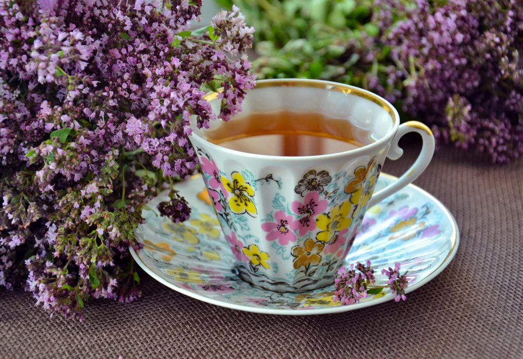 Auf einer beigen Tischdecke steht eine Tasse auf einer Untertasse.
Sie sind wellig geformt und haben rosane und gelbe Blüten drauf gemalt.
In der Tasse befindet sich eine hellbraune Flüssigkeit.
Um die Tasse herum ist lilaner Flieder drappiert.