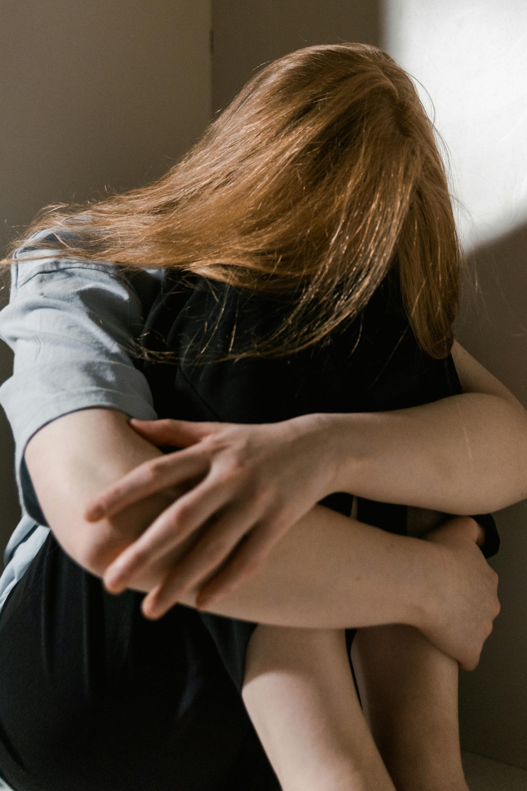 Eine Frau mit blonden Haaren sitzt in der Ecke eines Raumes.
Sie hat die Beine angezogen, die Arme drum herum und den Kopf gesenkt.