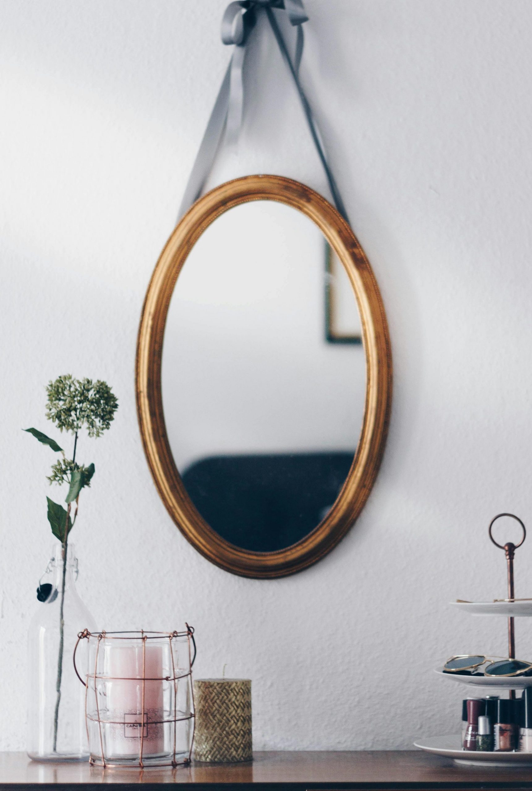Ein ovaler Spiegel hängt an einer weißen Wand.
Er hat einen goldenen Rahmen.
Unter ihm befindet sich ein Regal aus dunkelbraunem Holz.
Darauf steht eine Blume, zwei Kerzen und auf einer Etagere eine Sonnebrille und verschiedene Nagellacks.