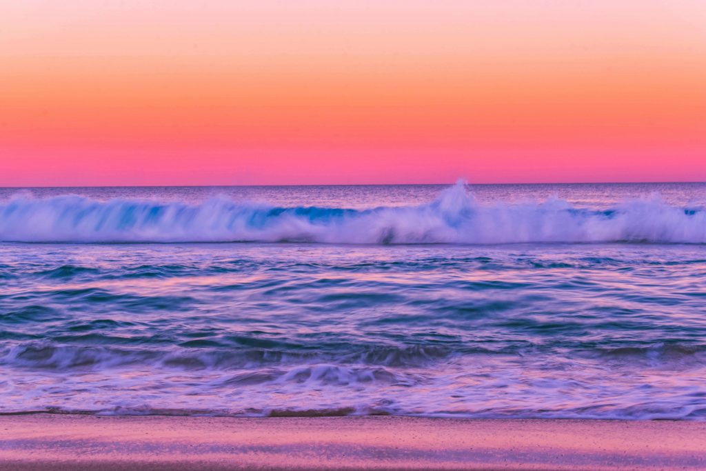 Strand, Meer und Himmel glänzen in den Farben apricot, orange, braun, rosa pink.
Eine türkisfarbene Welle sitzt auf dem Meer.