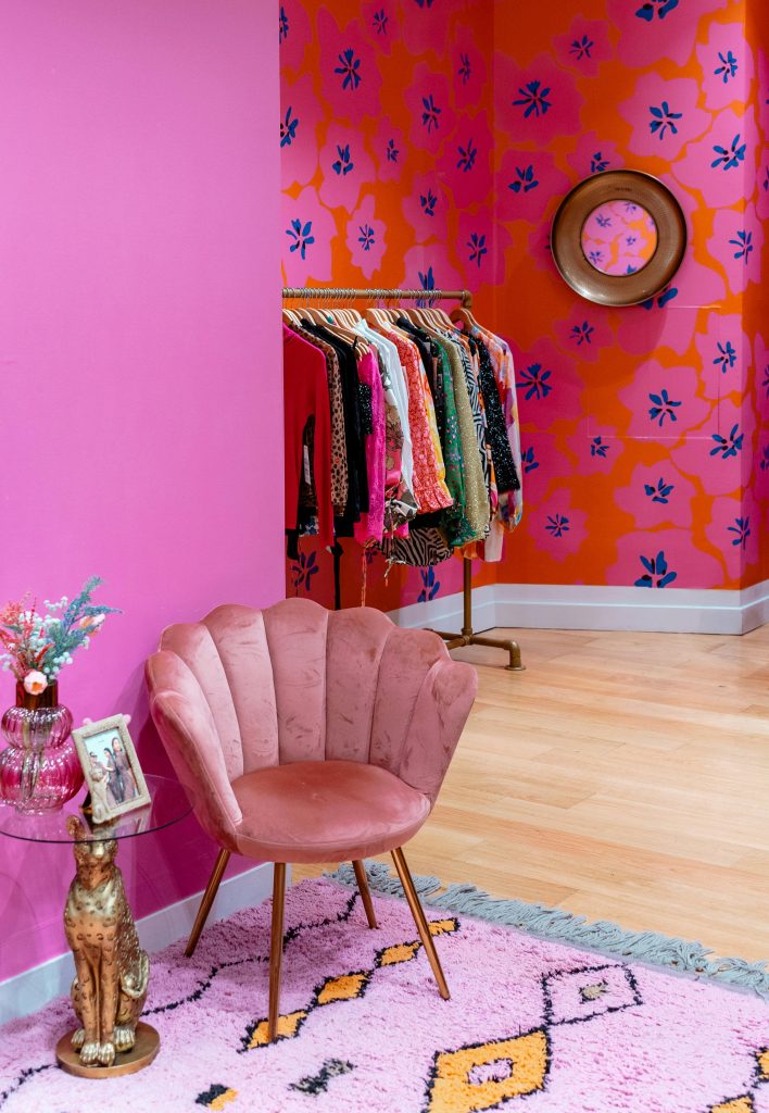Vor einer pink angestrichenen Wand steht ein altrosaner Polstersessel.
Er steht auf einem hellrosanen Teppich.
Daneben steht ein Glastischchen mit Blumen und einem Bilderrahmen.
Weiter hinten im Zimmer steht eine Stange mit diversen Kleidungsstücken auf Kleiderbügeln.