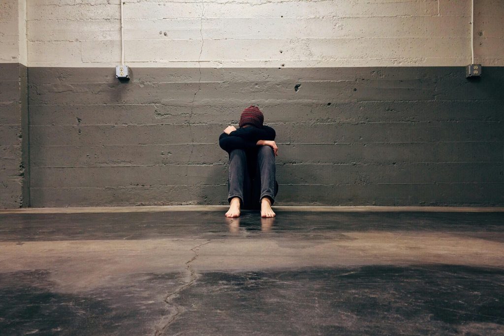 Auf einem kahlen, grauen Steinboden sitzt ein Mann.
Er lehnt an einer grauen Steinwand.
Er trägt eine dunkelrote Mütze, einen schwarzen Pulli, eine schwarze Jeans und ist barfuß.
Seine Knie sind angezogen, drauf liegen, verschränkt seine Arme und darauf sein Kopf.