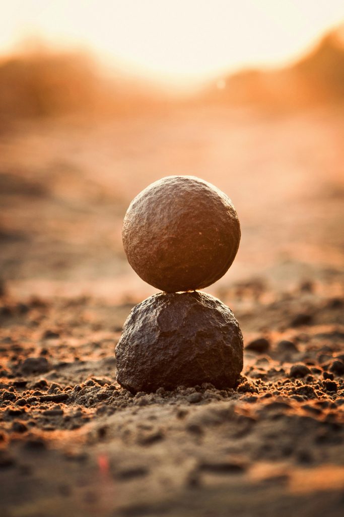Auf brauner Erde stehen zwei Moqui Marbles übereinander.
Sie werden umrahmt von goldenen Sonnenstrahlen, die von dahinter und leicht drüber kommen.
