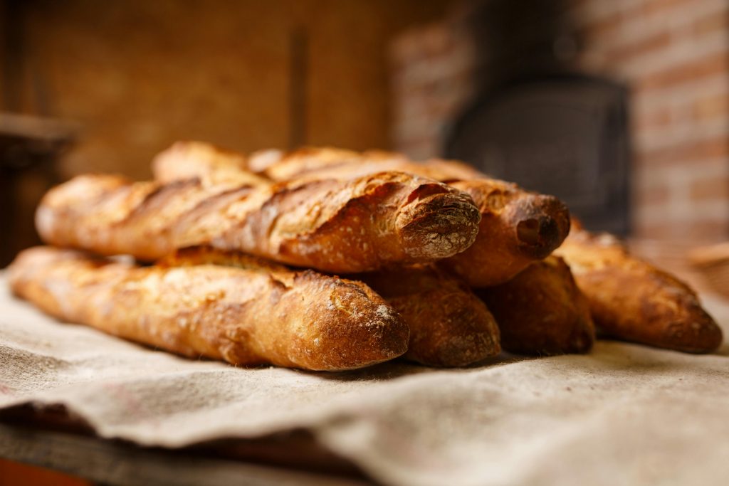 Auf einem Tisch liegt ein Tuch.
Darauf liegen sechs Stangen Baguette.
Im Hintergrund ist verschwommen eine Wand aus Ziegelsteinen und ein alter, schwarzer Backofen.
