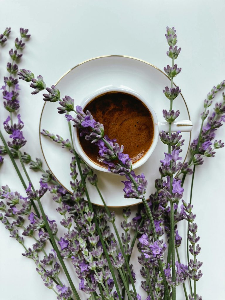 Auf einem weißen Untersetzer steht eine weiße Tasse. Darin befindet sich Kaffee.
Um sie Tasse und herum und auf ihr drauf liegen Stängel mit Lavendelblüten.