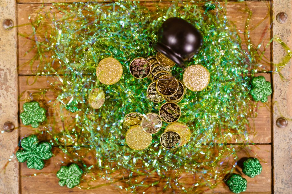 Auf einem braunen Tisch ist ein kleines, umgefallenes schwarzes Kesselchen.
Aus ihm sind ein paar Goldmünzen heraus gepurzelt.
Drum herum liegen grüne-glitzernde vierblättrige Kleeblätter.