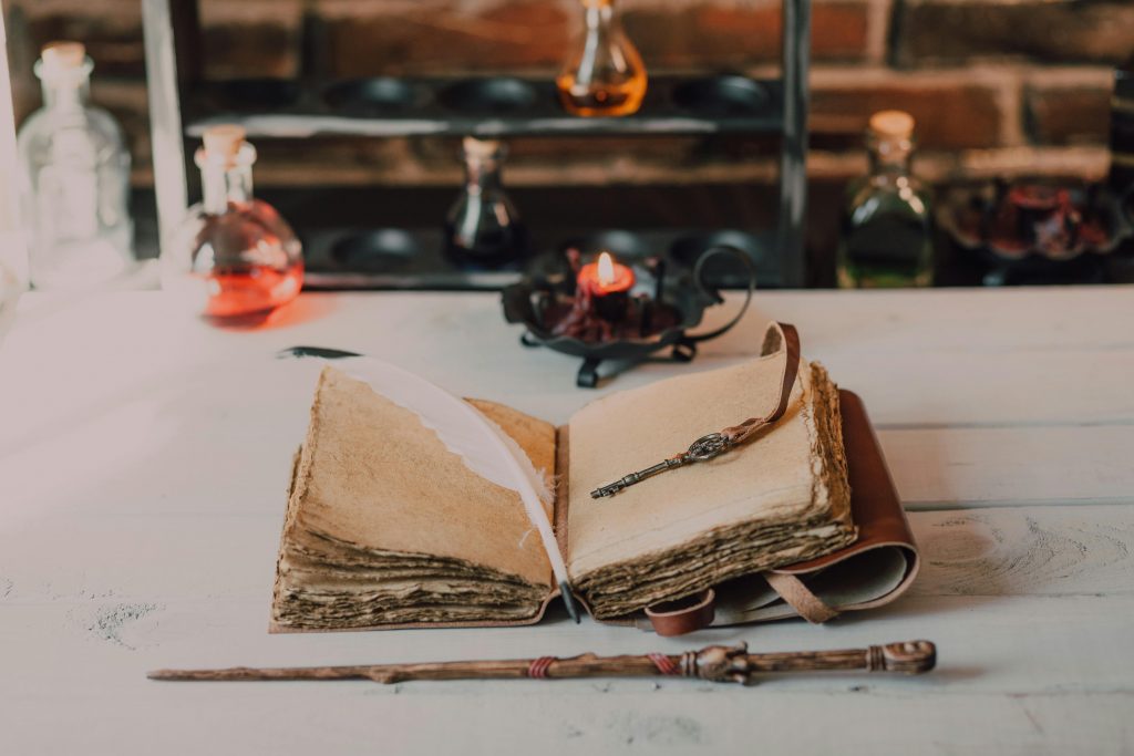 Auf einem alten Holztisch liegt ein altes Buch.
Es ist in der Bitte geöffnet.
Die Seiten sind abgegriffen und vergilbt.
Darauf liegt ein Schlüssel und eine weiße Feder.
Davor liegt ein Zauberstab,
Dahinter stehen Fläschchen mit verschiedenfarbigen Flüssigkeiten und eine Kerze.