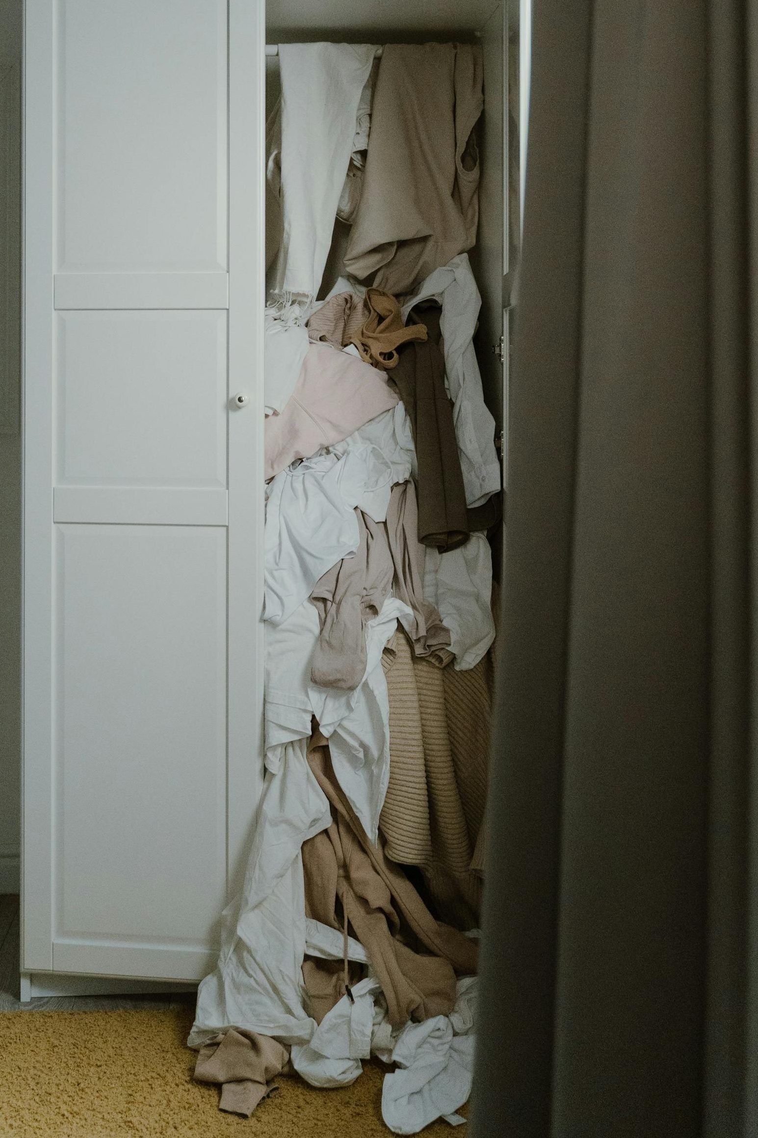Von einem weißen Kleiderschrank ist die rechte Tür geöffnet.
Daraus quillen viele verschiedene Kleidungsstücke hervor und bilden einen unübersichtlichen Haufen.