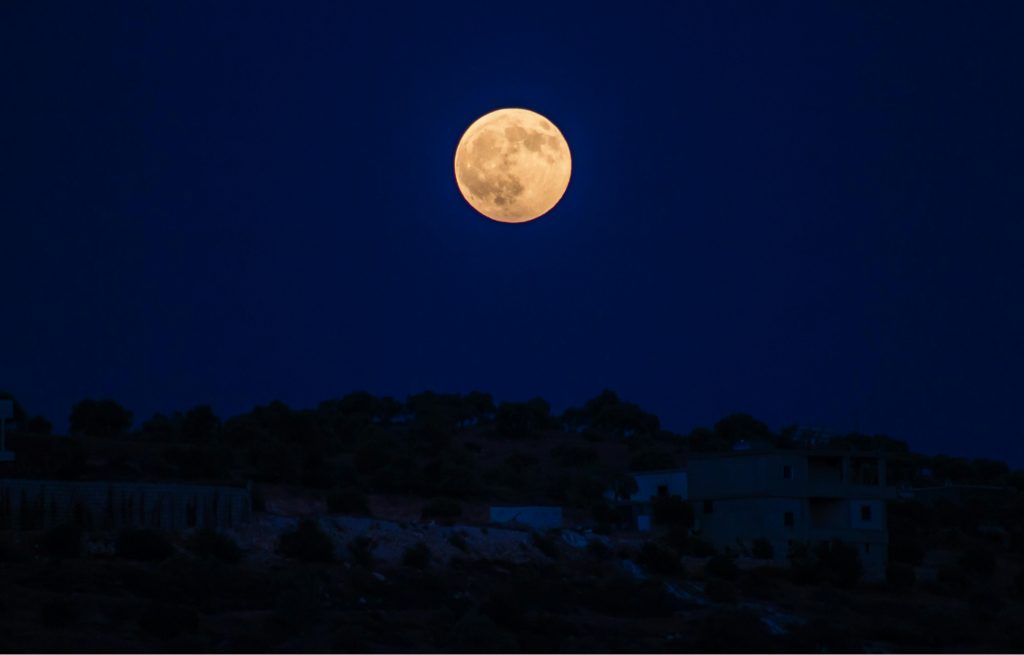 Vor einem dunkelblauen Abendhimmel steigt ein großer apricotleuchtender Vollmond empor.
Unter ihm befindet sich ein Hügel mit Bäumen und Häusern.