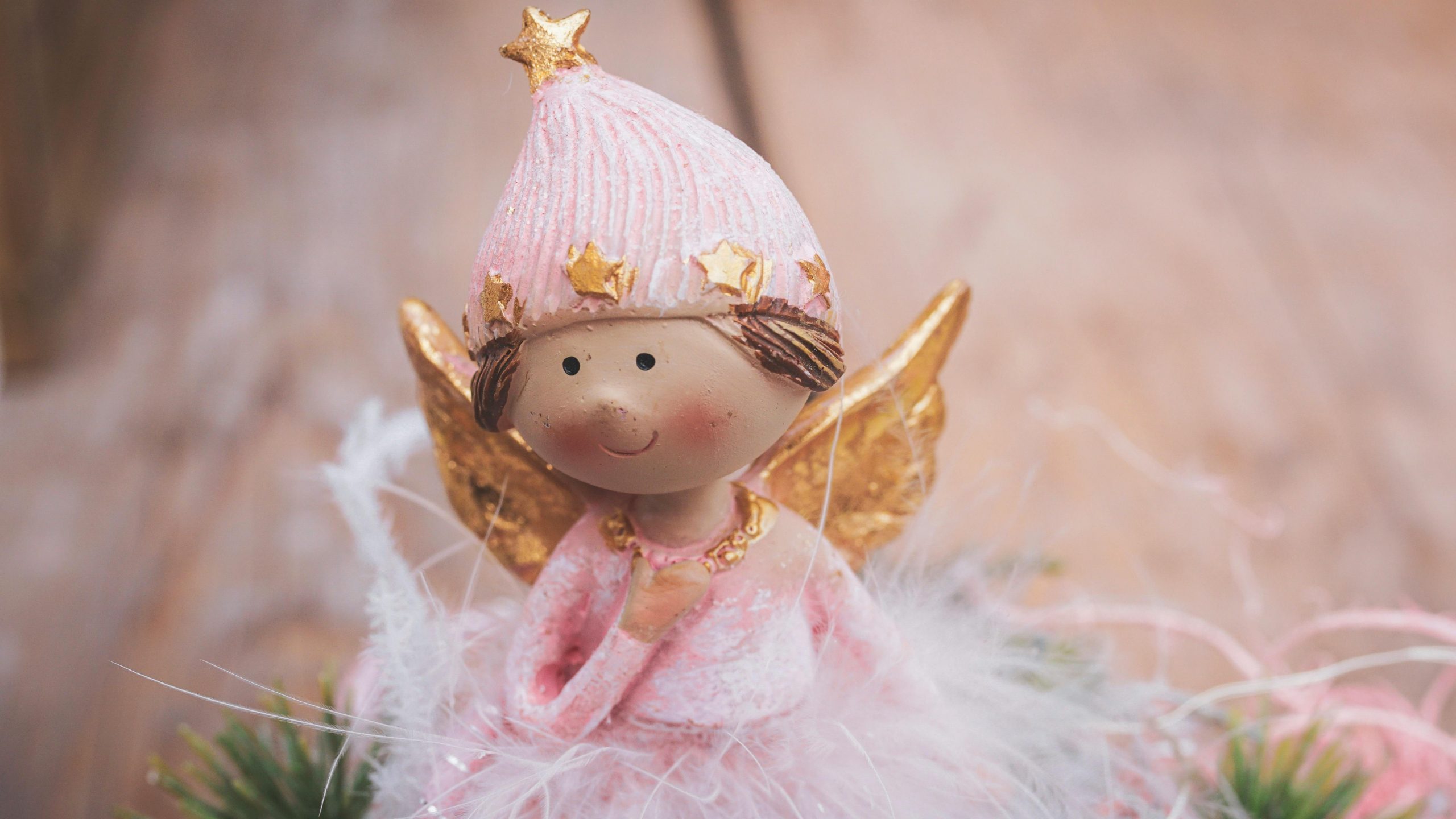 Auf einem braunen Holztisch steht die kleine Figur eines Engelchen.
Seine Haare sind braun und es hat eine rosa Mütze mit goldenen Sternchen auf dem Kopf.
Es trägt ein rosanes Kleidchen mit Federn und hat goldene Flügelchen.
Es lächelt süß und hält die rechte Hand auf die Brust.