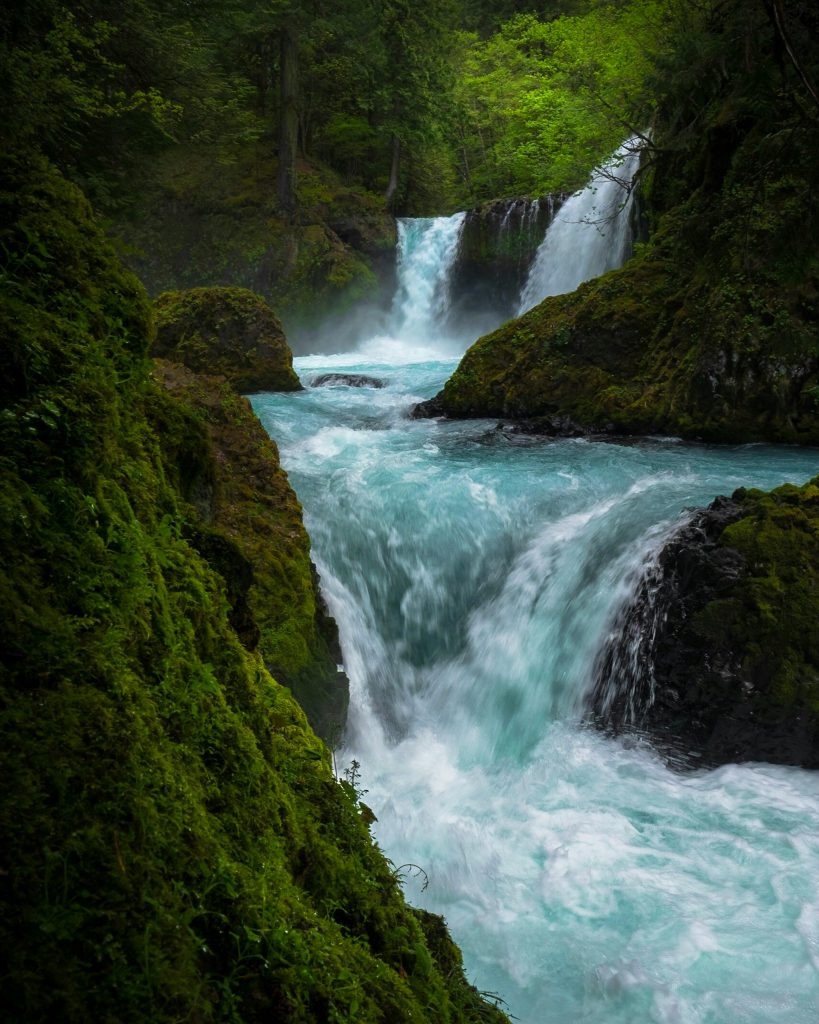 Durch Gestein, das von Moos grün gefärbt ist, schlängelt sich ein Fluss, der mit einem kleinen Wasserfall beginnt.